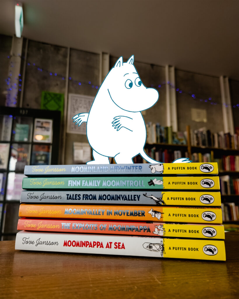 A cartoon Moomin stands on a pile of real "Moomin" books.